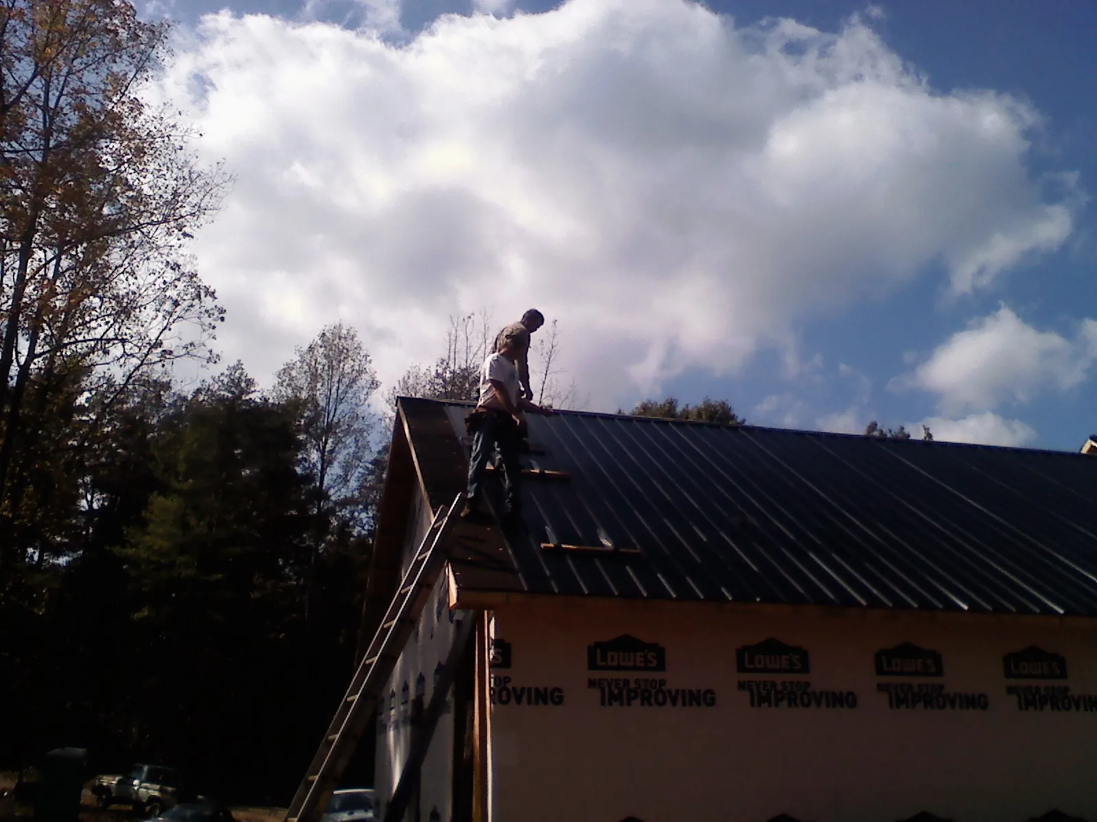 Roofer fastening dark metal panels high against a cloudy sky, from a Roof Inspection job in Yantic